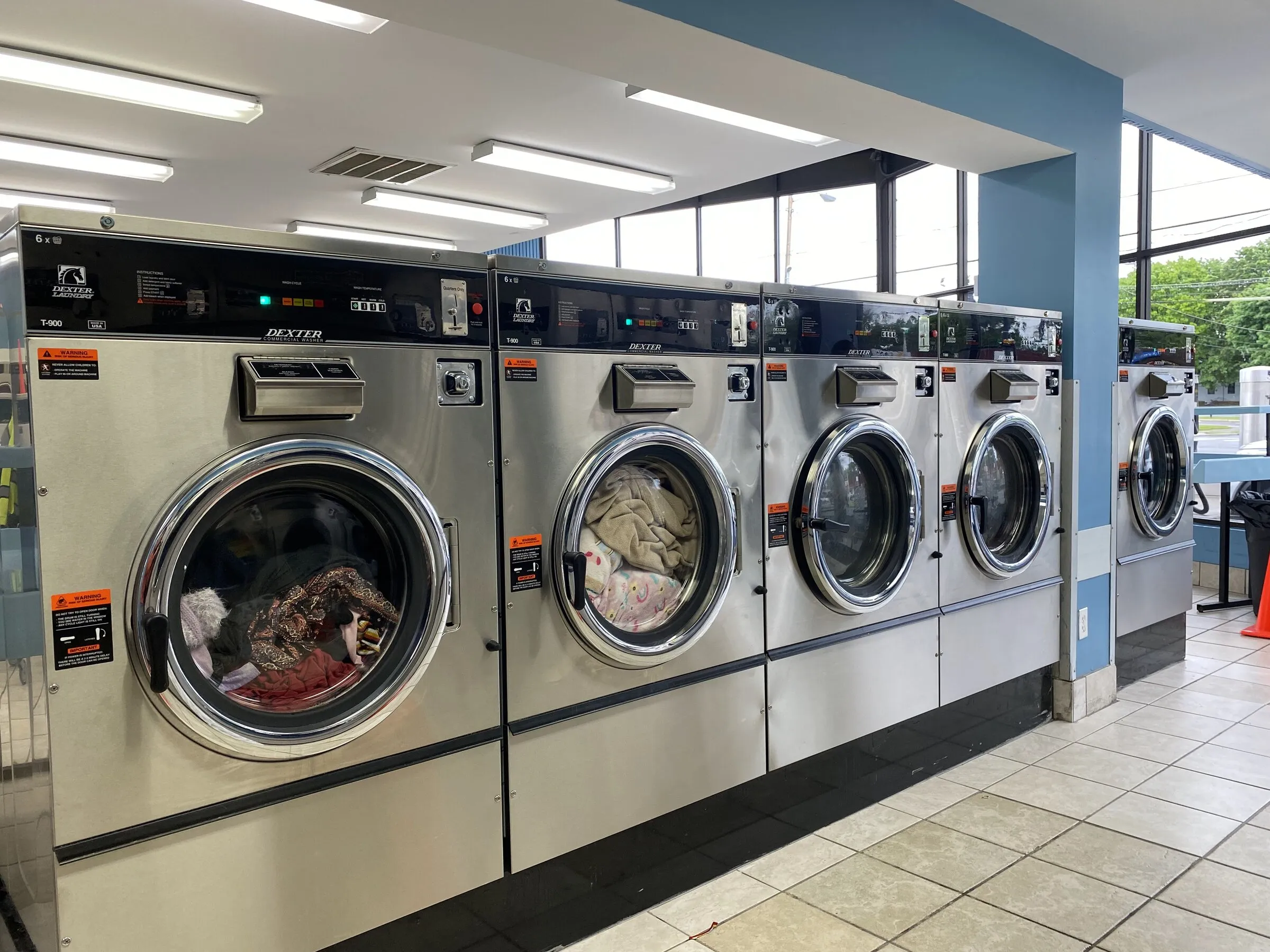Five 60-pound washers in a row, three of them running with clothes and a comforter inside, daylight from the windows behind.