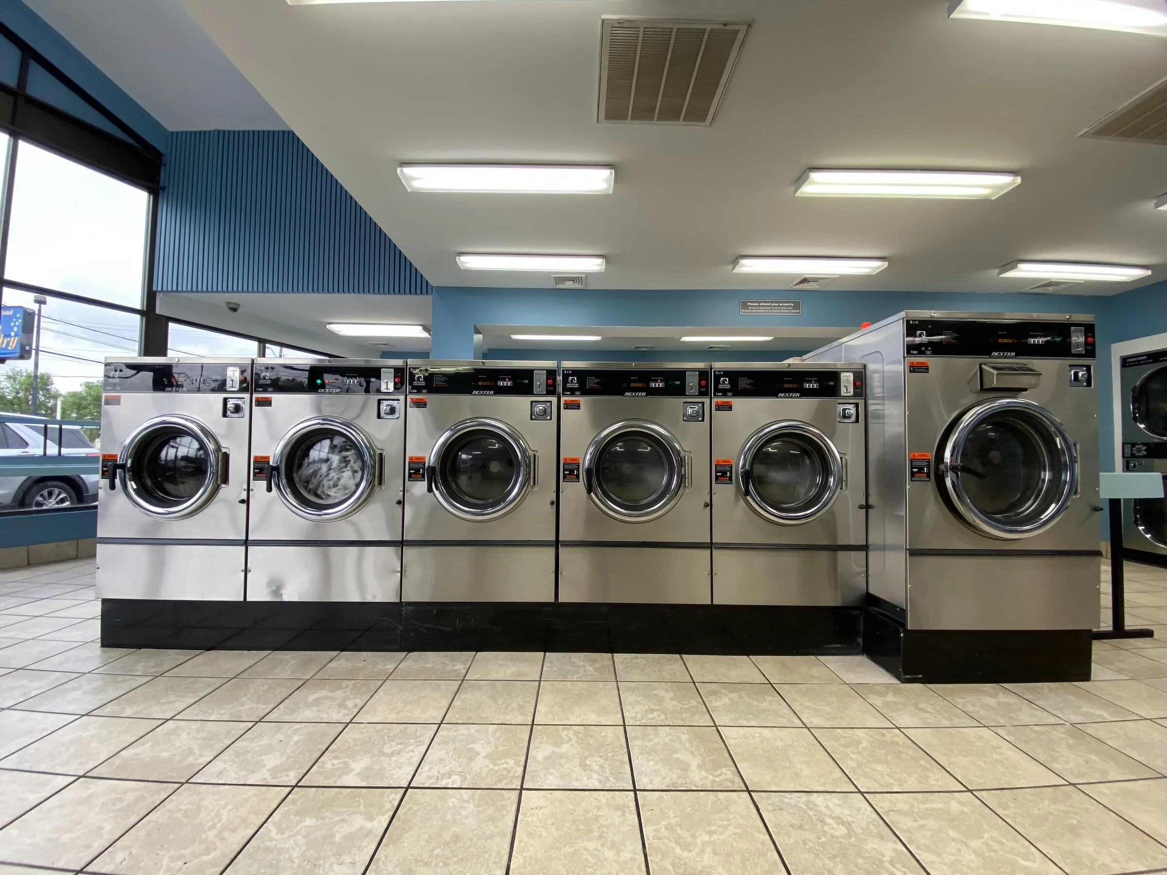 A row of front-load washers in bright daylight, with the parking lot visible through the windows.
